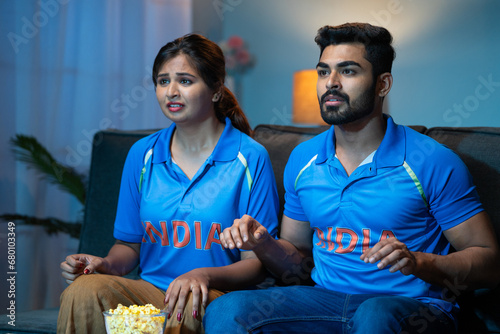 shocked Young couples while watching live cricket match on tv or television at home - concept of anticipation, sports fever and entertainment