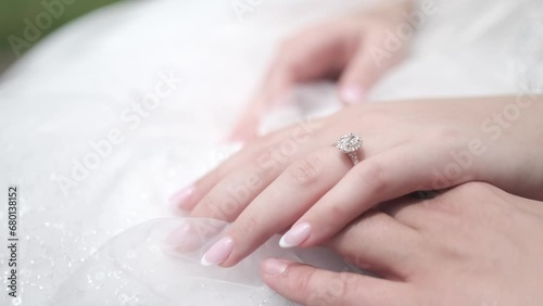 A 4k close up shot of a brides hand wearing a sparkly wedding ring. The bride is wearing a white wedding dress and  her nails have a french manicure. Her hand is resting on top of the grooms. photo