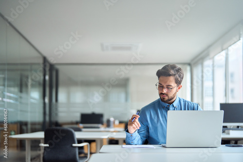 Busy young latin businessman working in office using laptop checking documents. Serious professional business man hr manager analyzing accounting report doing project overview. Copy space. photo