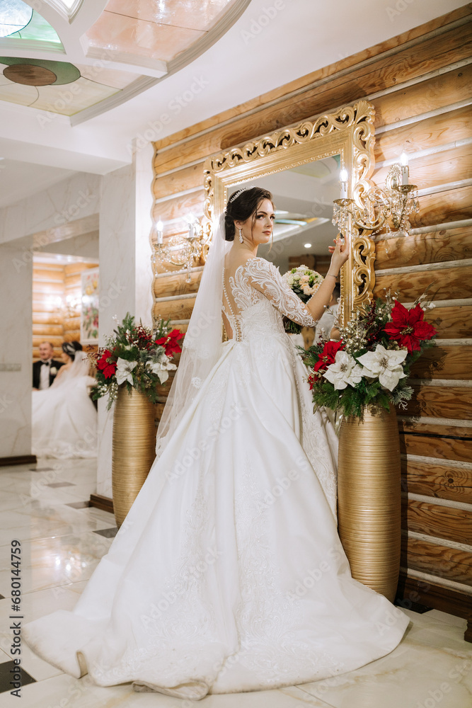 Fototapeta premium Elegant brunette bride poses in a white dress near the mirror. Portrait of the bride, wedding makeup and hairstyle, wedding fashion. Beautiful bride in a veil.