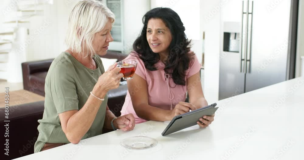 Happy diverse senior women using tablet and drinking tea in sunny kitchen, slow motion