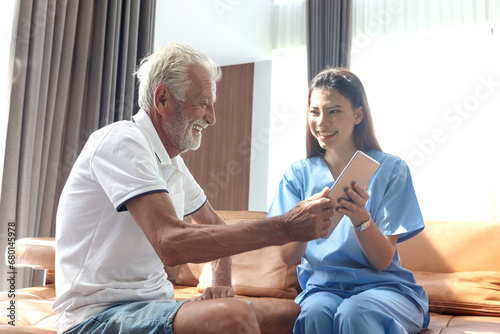 Female nurse takes care of senior patient and teaches him to use digital tablet at living room house, elderly mature grandfather learns to online internet and high-tech, nursing medical health care.