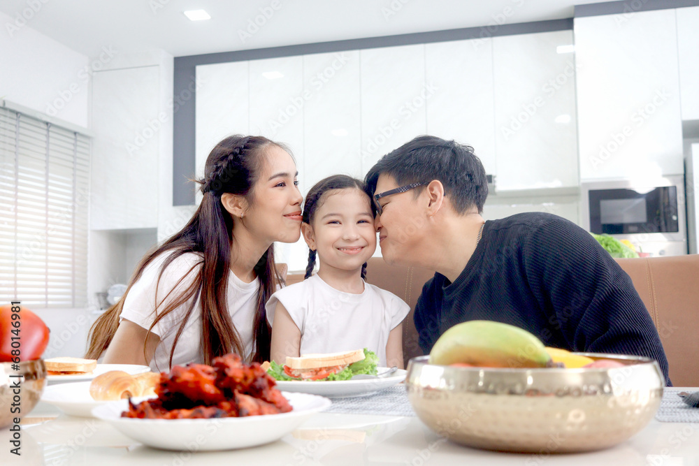 Happy family has meal in dining room. Mother father kid daughter sit at dining table, have fun during breakfast, parent kiss girl child at dining table. Family enjoy eating food together. Happy family has meal in dining room. Mother father kid daughter sit at dining table, have fun during breakfast, parent kiss girl child at dining table. Family enjoy eating food together.