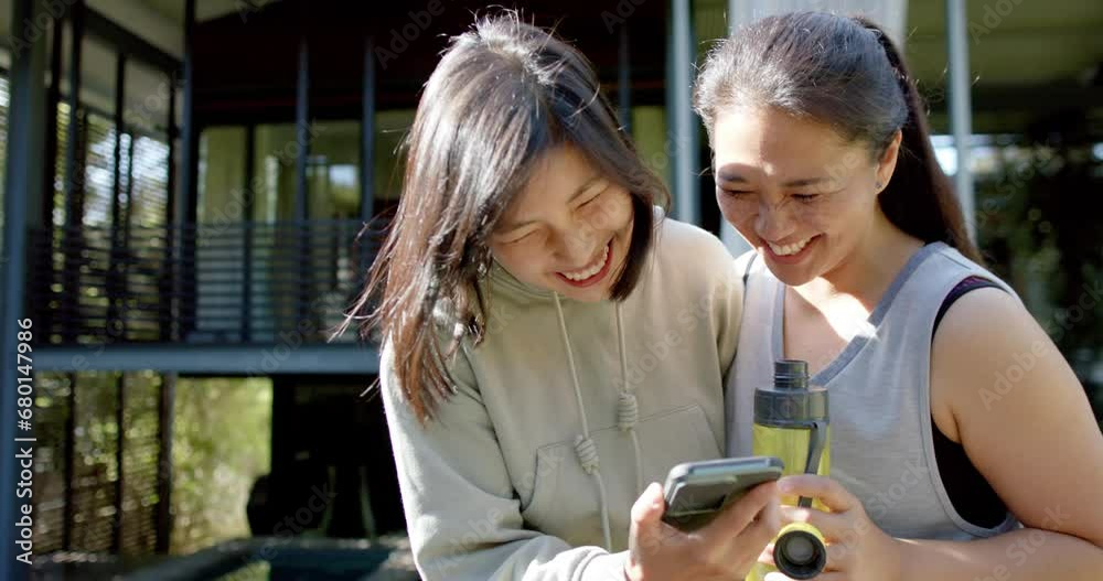 Happy asian female friends with water bottle and smartphone laughing on terrace, slow motion Happy asian female friends with water bottle and smartphone laughing on terrace, slow motion