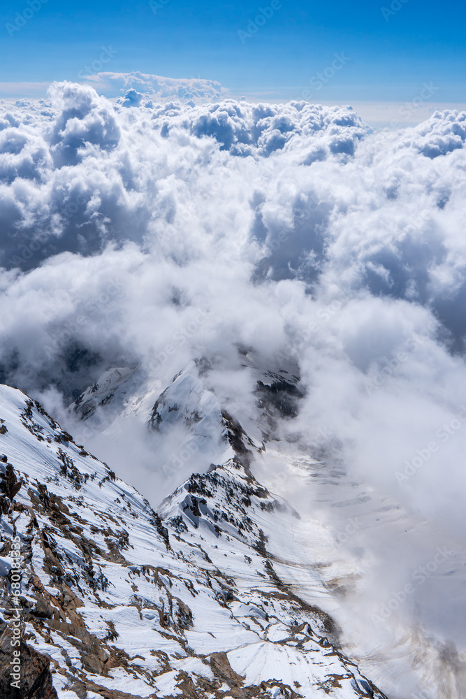 alpine landscape with peaks covered by snow and clouds Cloudy mountains. Mountains in clouds at sunrise in summer. Aerial view of mountain peak with green trees in fog. Top view of mountain valley 