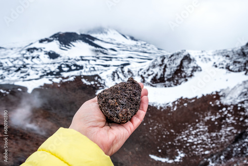 Tourist hand holding lava stone on Mount Etna crater, Sicily, Italy. Warm volcanic lava rock formation. Landscape of craters slopes covered with snow, clouds and smoke photo