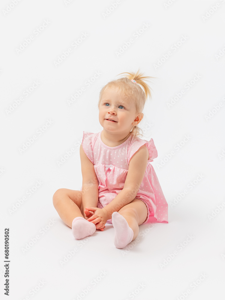 A cute blonde girl 1-2 years old in a pink dress is sitting on the floor and enthusiastically looking at something on a white background. Copy space. A cute blonde girl 1-2 years old in a pink dress is sitting on the floor and enthusiastically looking at something on a white background. Copy space.