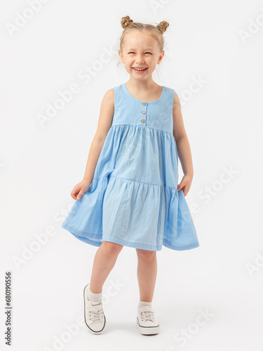 A cute Caucasian 5-year-old girl with a beautiful smile with a fashionable hairstyle stands on a white background in full height in a blue summer sundress and laughs with narrowed eyes. photo