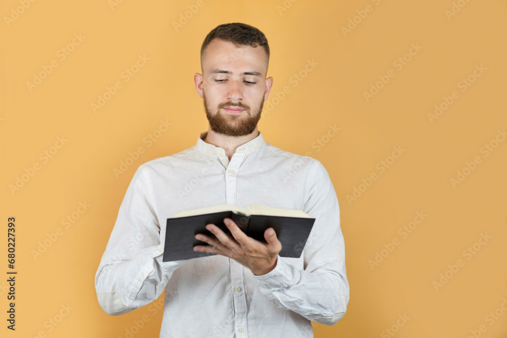 Black hardcover book in the hands of a young guy with a beard on a yellow background, the guy is enthusiastically reading and memorizing Bible verses Black hardcover book in the hands of a young guy with a beard on a yellow background, the guy is enthusiastically reading and memorizing Bible verses