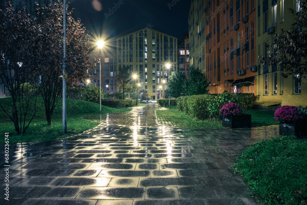 Fototapeta premium Night park paths and colored houses on a rainy autumn night. Night paths, benches, lanterns in a beautiful residential complex. Night autumn park with lanterns, benches, yellow leaves. Kyiv. Ukraine