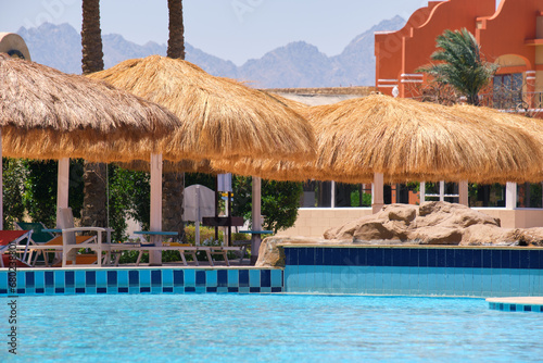 Empty deck chairs under straw shade umbrellas on swimming pool side in tropical resort. Summer vacations and getaway concept