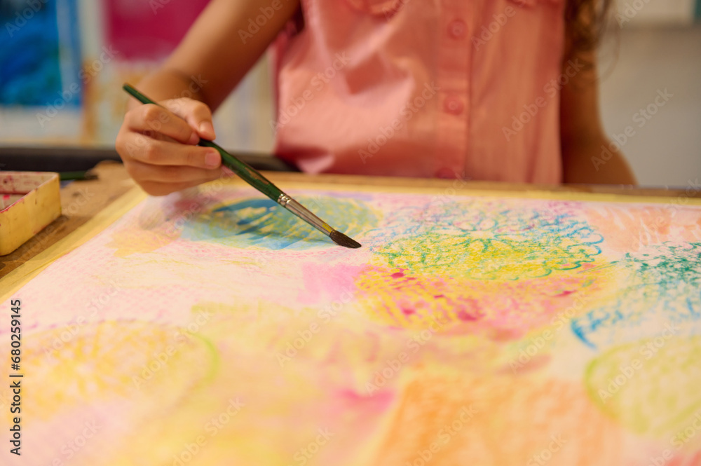 Close-up hand of little artist sitting with painting accessories, holding paintbrush and drawing her unfinished painting Close-up hand of little artist sitting with painting accessories, holding paintbrush and drawing her unfinished painting
