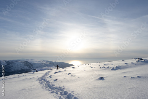 A tourist walks along a snowy mountain slope. Footprints in the snow. There is a solar halo in the sky above the sea. Traveling and hiking in Siberia and the Russian Far East. Magadan region, Russia. © Andrei Stepanov