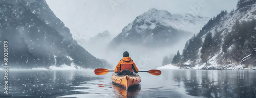 Man floats on a kayak in mountain lake on a snowy cold winter day. Rear view of male paddling canoe. Fog and snow fall on the water on the background. Copy space panorama.