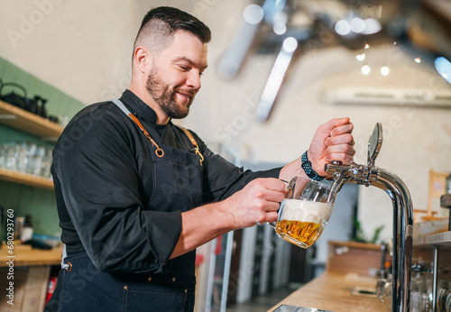 Smiling stylish bearded barman dressed black uniform with an apron tapping fresh lager beer into glass mug at bar counter. Successful people, beer consumption, beverages industry concept image