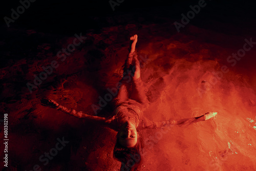 Ethereal woman floats tranquilly in lagoon illuminated by crimson light, nighttime serenity enveloping her form. Feminine grace drifts gently, waterscape aglow in ruby tones providing surreal setting. photo