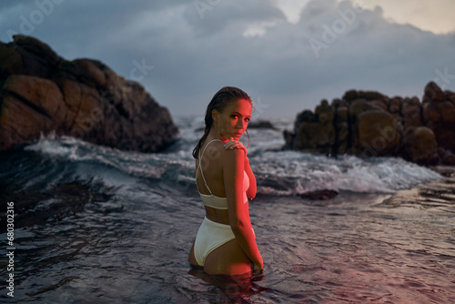 Attractive young woman enjoys nocturnal swim in ocean, illuminated by crimson light. Female beauty in stylish swimwear, night-time leisure at sea, tranquil waterscape surrounds.