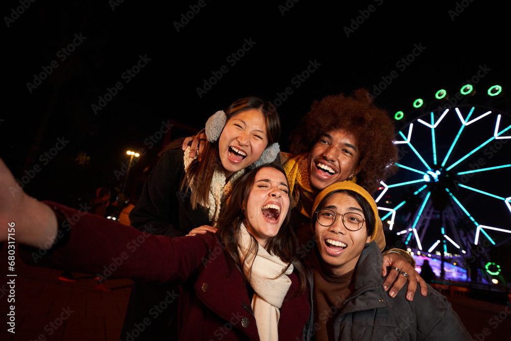 Selfie phone multiracial group of happy colleagues having fun in winter. Young smiling generation z people excited together enjoy community. Surprised face of friends at amusement park at night. 