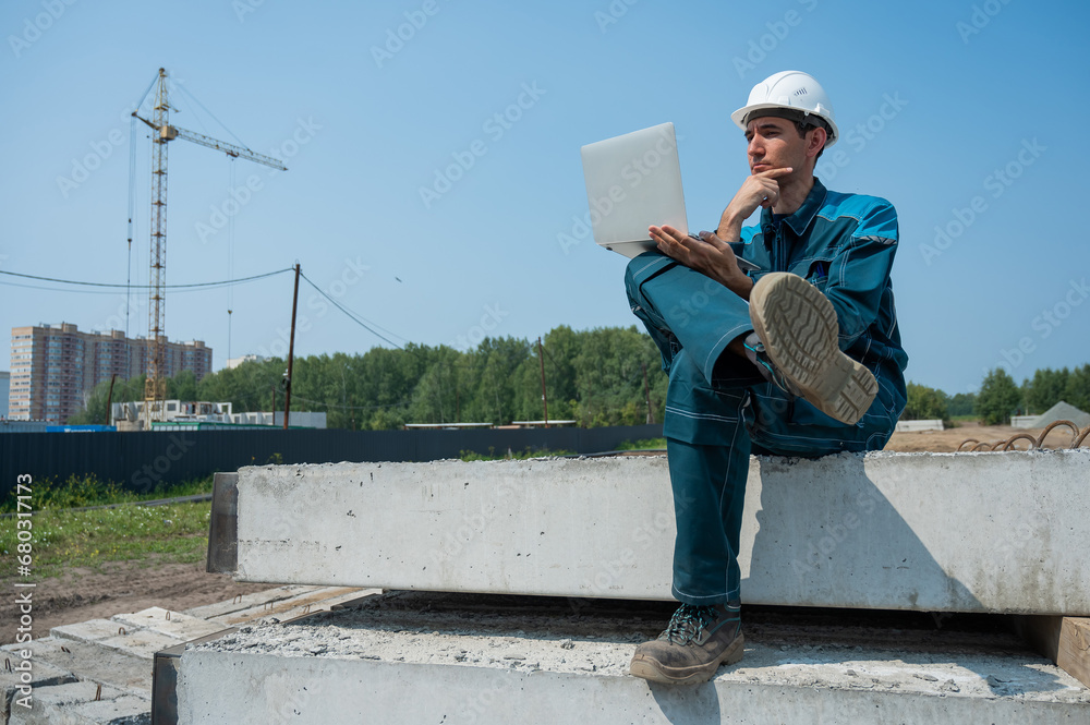 Caucasian male builder in hardhat sits on floor slabs and uses laptop at construction site. Caucasian male builder in hardhat sits on floor slabs and uses laptop at construction site.