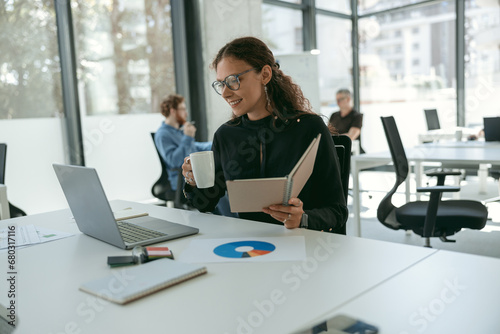 Smiling female entrepreneur working on laptop and drinking coffee while sitting in modern coworking