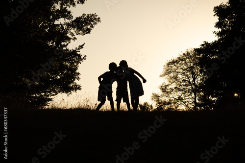 Black shadow silhouette of three kids standing together in the forest  holding each other on a sunset sky and trees background. Friendship or happy summer adventure concept photo.