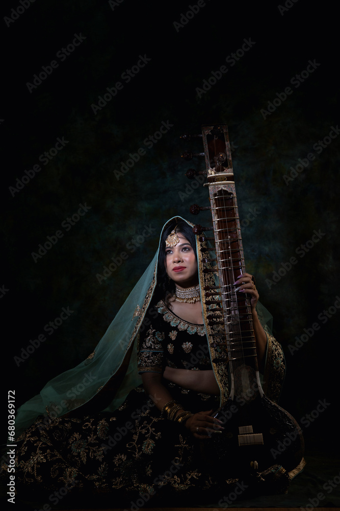 Obraz premium beautiful indian woman in brown bridal lehenga with green dupatta over head and heavy jewellery and bangles sitting on a chair in luxury background holding veena ( harp)