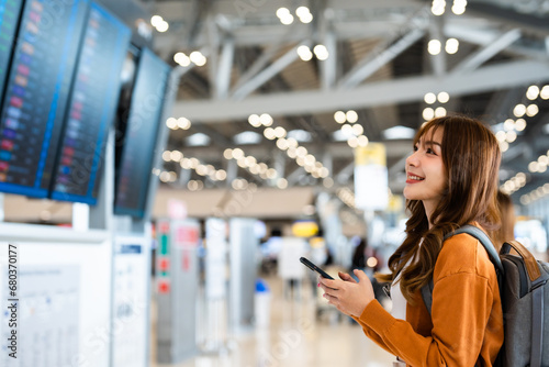 Young asian woman in international airport  using mobile smartphone and checking flight at the flight information board