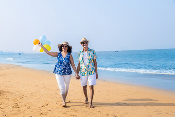 Retired Senior indian couple with colorful balloon in hands walking together at  beach. Enjoying vacations holidays at beach.  Love, romantic, bonding, relationship, fun, Copy space.