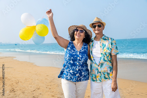 Senior indian couple with colorful balloon in hands walking together at the beach. Enjoying vacation, holiday at beach. Copy space. Retirement life.