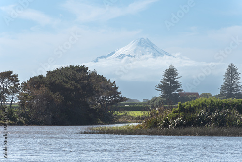 Distant view of snow-capped Mount Taranaki peeking through low clouds with a rural landscape and Lake Lowell's in the foreground.