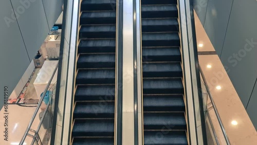 Escalators going up and down in the mall. The escalator is located in the middle of the mall, and is surrounded by restaurants. Escalators are made of metal and glass, clean and well maintained