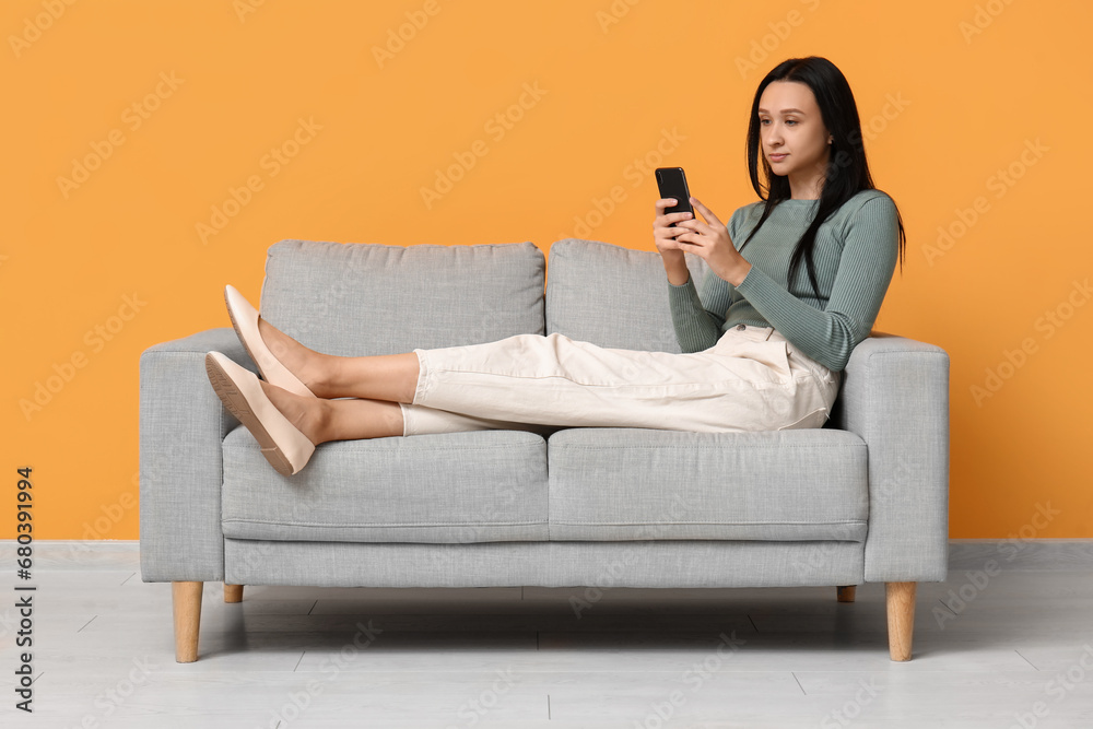 Obraz premium Beautiful young woman with mobile phone sitting on comfortable sofa near yellow wall