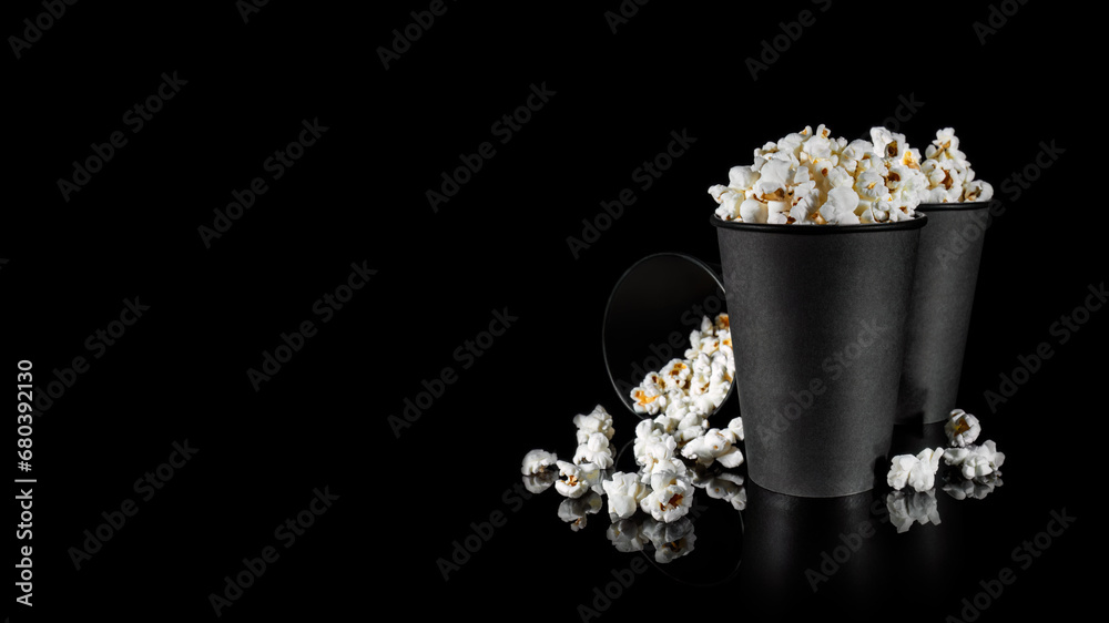Two black standing paper cups with popcorn and a third next to spilled popcorn on a black background with reflection. Banner. Copy space. Two black standing paper cups with popcorn and a third next to spilled popcorn on a black background with reflection. Banner. Copy space.