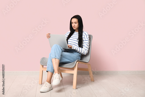 Beautiful young woman with laptop sitting on comfortable chair near pink wall