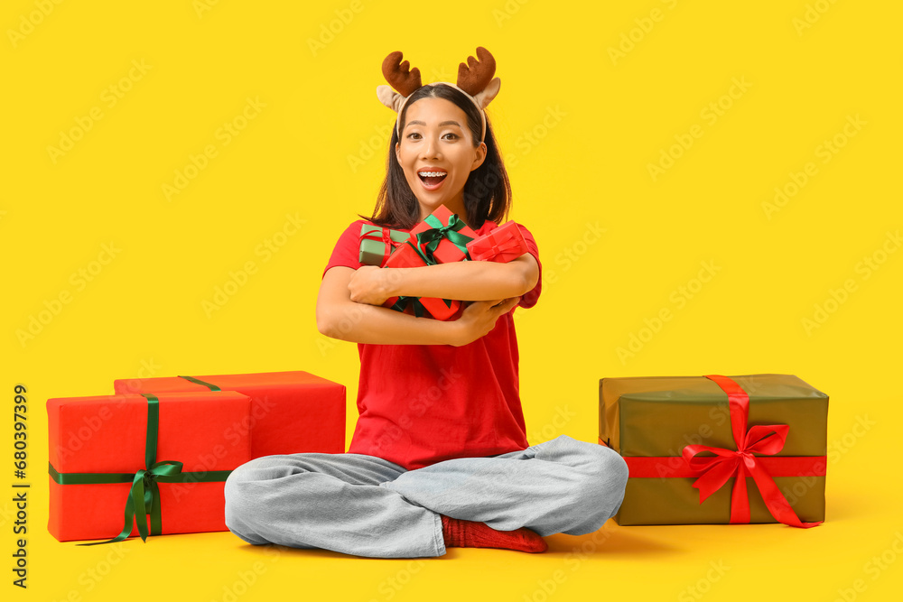 Surprised young Asian woman in reindeer horns with Christmas gift boxes on yellow background