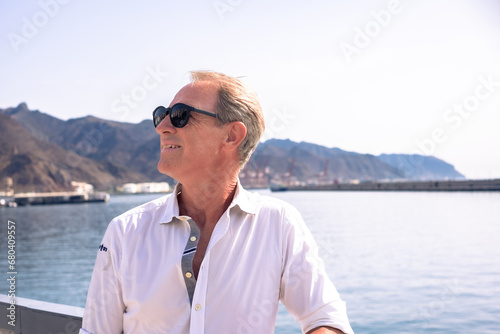 Portrait of handsome middle aged man with sunglasses at sea enjoying outdoors and vacation. Smiling relaxed people in white shirt looking away