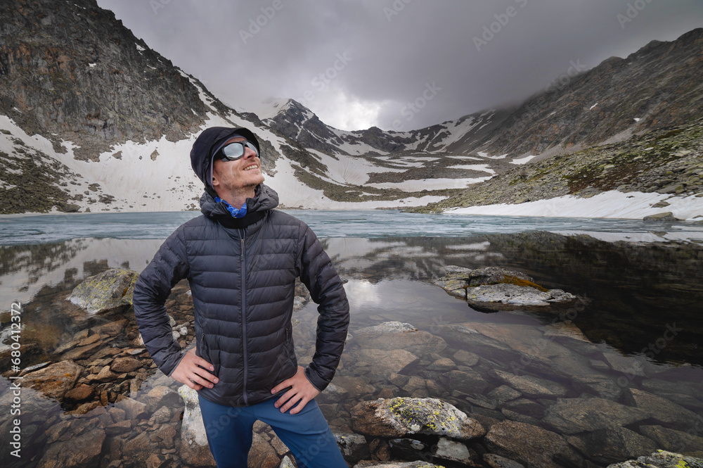 young tourist stands on the top of a mountain against the backdrop of a mountain lake. Happy guy smiling at the camera. Tourism, sport and travel concept