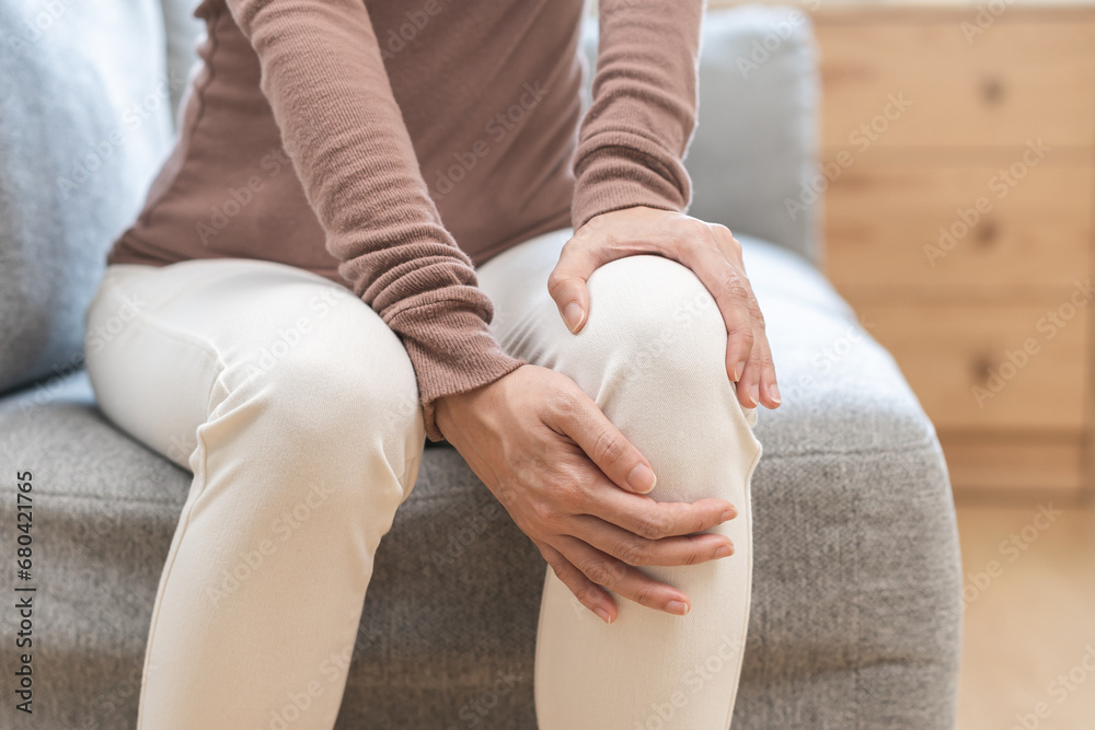 Health care of knee pain concept, close up hand of woman arthritis sitting joint ache, sore cramp or sprain tendon in leg on sofa at home. Osteoarthritis chronic disease of body problem from trauma. - obrazy, fototapety, plakaty 
