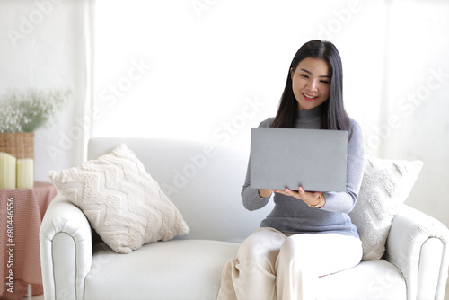 Young asian woman using computer laptop while seated on couch at home. Happy girl relaxing comfortable, using laptop in the living room, spending lazy weekend, watching movie, shopping online.