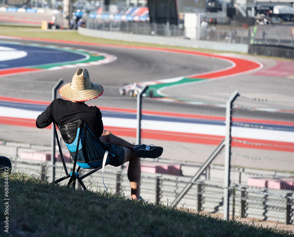 Rear view of lone spectator, wearing a sun hat, looking at a racing car on track.