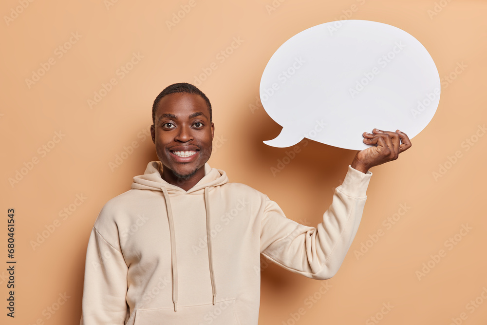 Waist up shot of handsome dark skinned young man with beard happy toothy smile on face holds blank speech bubble for your ad or promotion wears casual sweatshirt isolated over brown background Waist up shot of handsome dark skinned young man with beard happy toothy smile on face holds blank speech bubble for your ad or promotion wears casual sweatshirt isolated over brown background