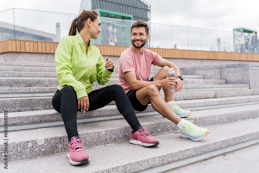 A fitness trainer and a client communicate during a break, friends train together in sportswear. Uses sportswear and accessories app in smart watch, male and female runner.