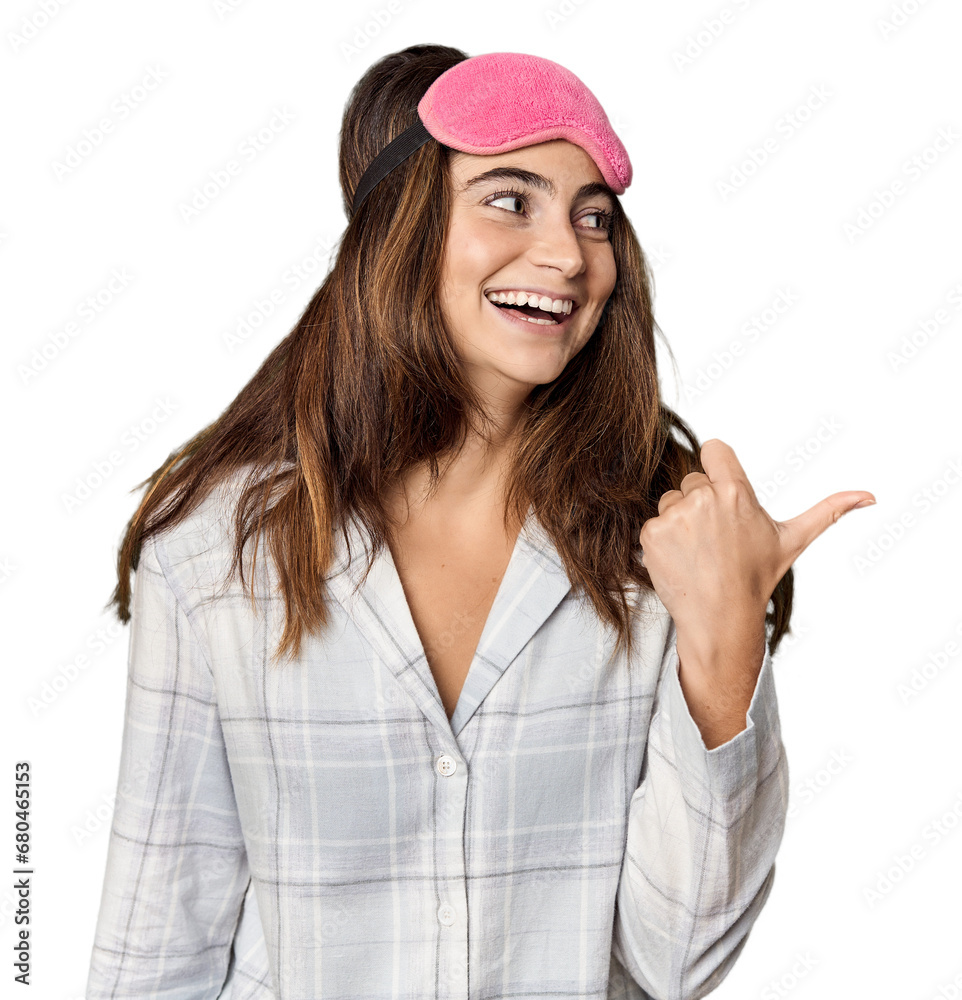 Young Caucasian woman in pajamas with mask points with thumb finger away, laughing and carefree. Young Caucasian woman in pajamas with mask points with thumb finger away, laughing and carefree.
