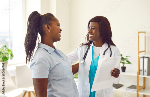 Portrait of smiling friendly female african american doctor therapist talking with a young woman patient holding report file with appointment standing in office. Medicine and health care concept. photo