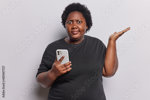 Confused overweight curly woman shrugs shoulders holds smartphone has connection error annoyed with discharged or broken cellphone dressed in casual black t shirt isolated over white background photo