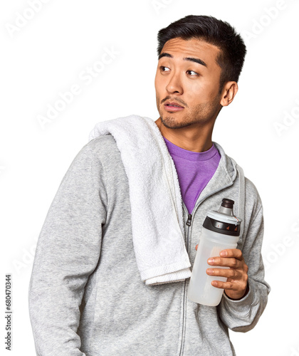 Chinese man with sportswear, towel, bottle in studio looks aside smiling, cheerful and pleasant.