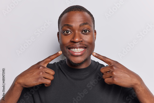 Young positive dark skinned man points at toothy smile shows perfect teeth dressed in casual black t shirt attracts your attention to mouth dressed in casual black t shirt isolated on white background