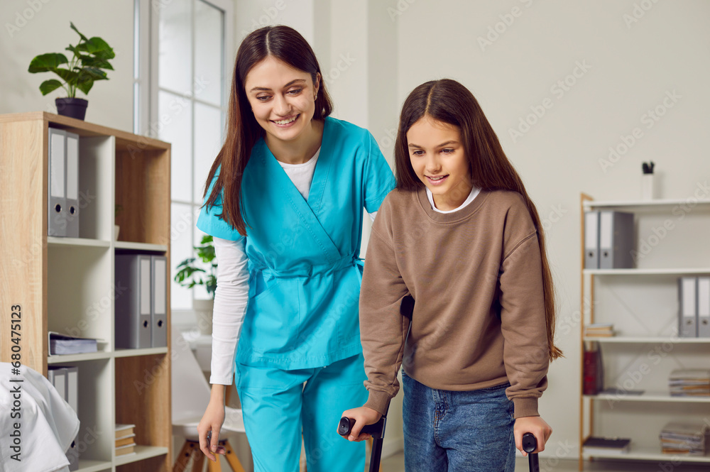 Portrait of a young cheerful brunette nurse helping a child girl patient to walk with her crutches. Friendly physiotherapist or paediatrician doctor helping disabled female teenager in rehab Portrait of a young cheerful brunette nurse helping a child girl patient to walk with her crutches. Friendly physiotherapist or paediatrician doctor helping disabled female teenager in rehab
