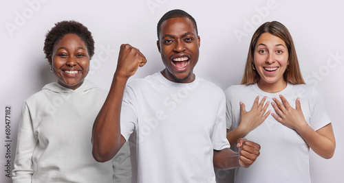 Dark skinned bearded man triumphant exclaims loudly clenches fist celebrate something stands in centre between two cheerful women smile happily dressed casually isolated over white background photo