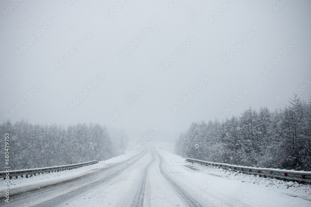 Photo of winter road during the snowfall in Magadan, Russia. Snow showers on trees and hills. Fog and haze, low visibility due to snowstorm. Extreme weather conditions. Car approaching in the distance Photo of winter road during the snowfall in Magadan, Russia. Snow showers on trees and hills. Fog and haze, low visibility due to snowstorm. Extreme weather conditions. Car approaching in the distance